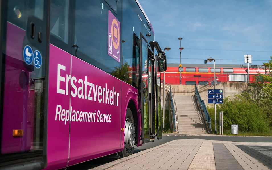 Zu sehen ist ein violetter Bus mit der Aufschrift "Ersatzverkehr" am Bahnhof, im Hintergrund sieht man einen roten Zug.