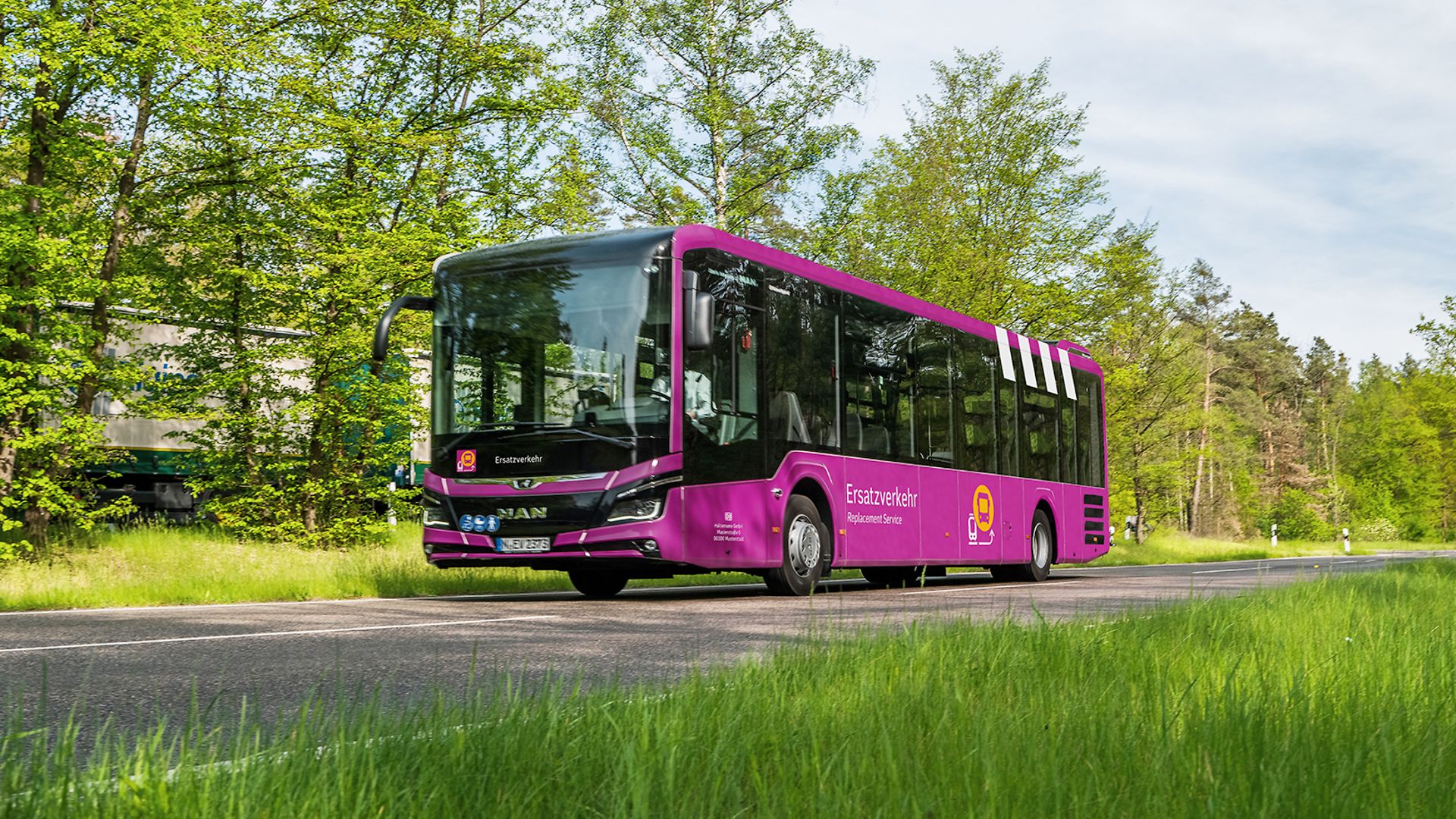 Ein violetter Ersatzverkehr-Bus fährt auf der Landstraße, umgeben von Bäumen und Wiese.